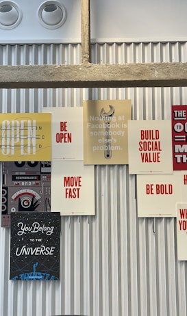 A collection of motivational and inspirational posters arranged on a corrugated metal wall. The posters feature phrases such as 'BE OPEN', 'MOVE FAST', 'BE BOLD', and 'BUILD SOCIAL VALUE' in bold typography. The display includes a stencil-style graphic of a wrench and other elements that convey a tech and innovation theme.