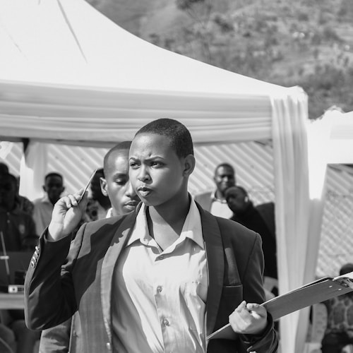 A person in business attire is holding a pen and a clipboard, walking purposefully with a focused expression. In the background, there is a gathering of people and a tent set up outdoors.