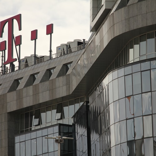 A modern office building with a glass facade features sharp, angular architectural lines. A large red 'T' logo is prominently displayed on the rooftop, accompanied by several antenna structures.